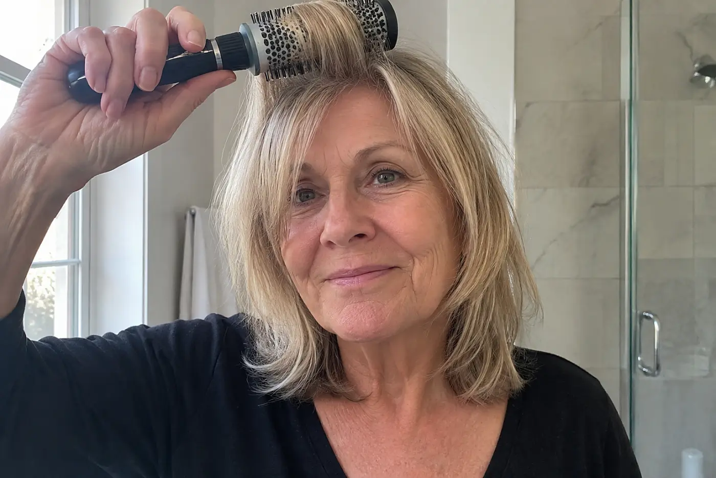 A woman guides her curtain bangs with a round brush in her bathroom.