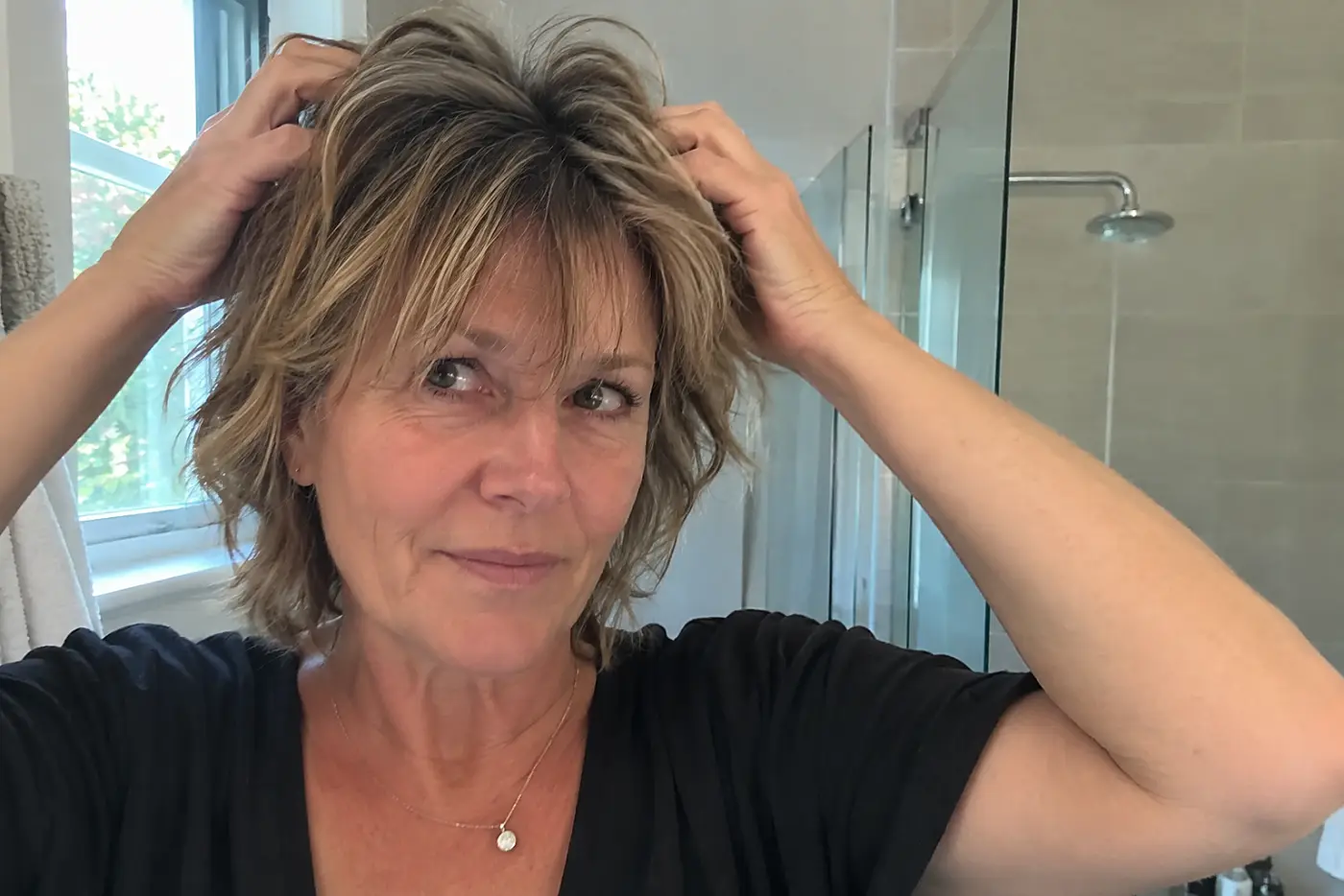 A woman shakes out the crown of her mid-neck shag in a bathroom mirror.