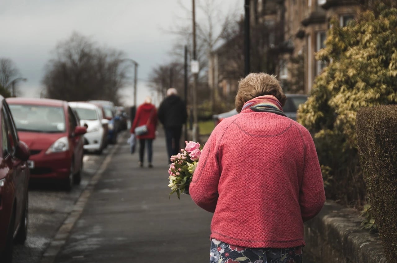 Older woman in a pink jacket carrying flowers walking along a residential sidewalk with parked cars