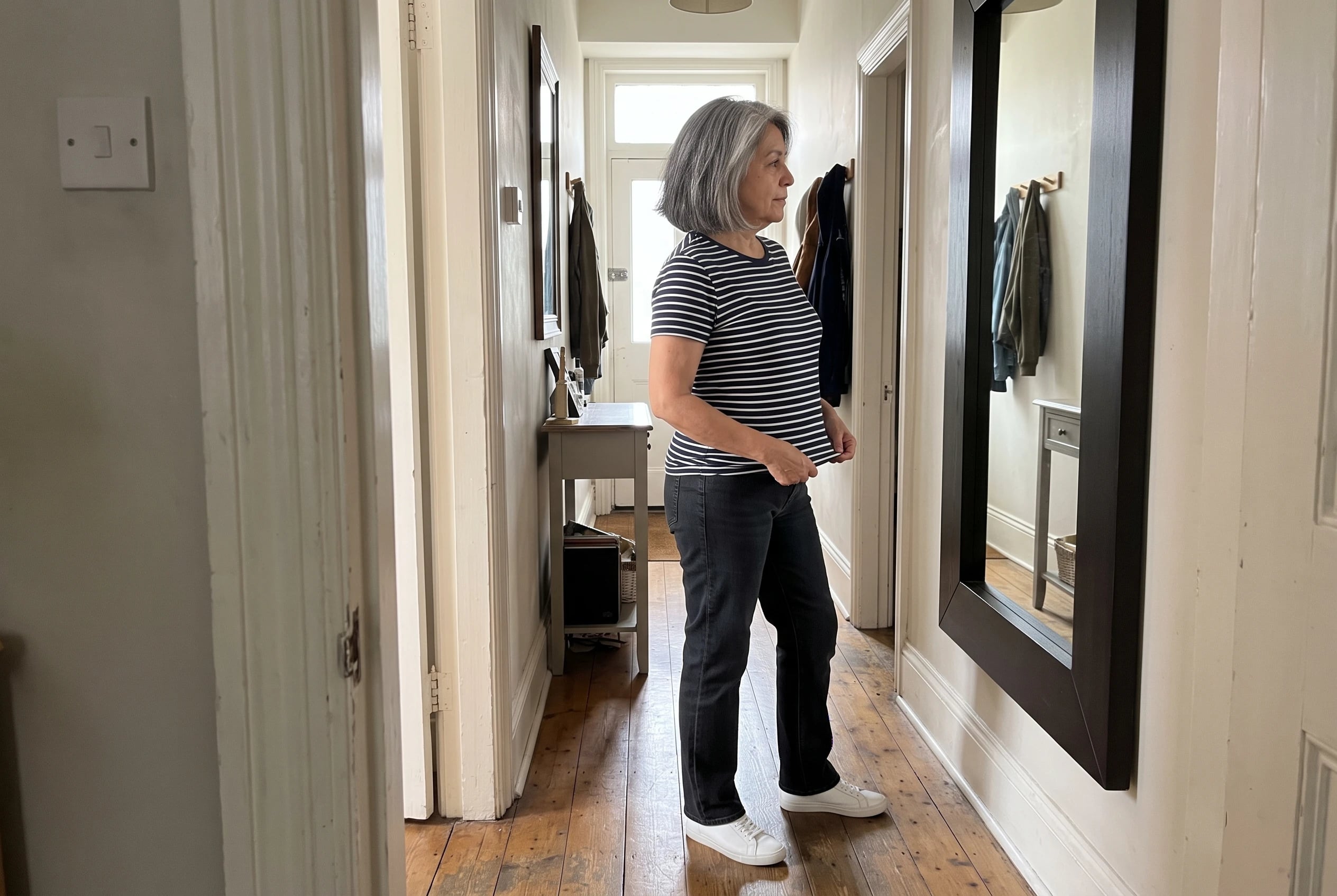 A woman in her sixties wearing a striped tee and dark jeans looks in a mirror.