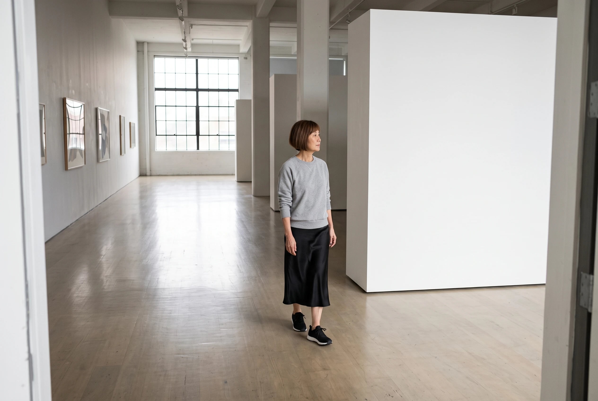 A woman in her sixties walks through a gallery wearing a gray sweatshirt, satin skirt, and trainers.