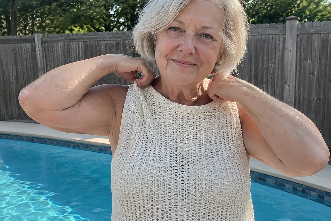 Older woman pulling a sleeveless knit cover-up over her swimsuit beside a pool.