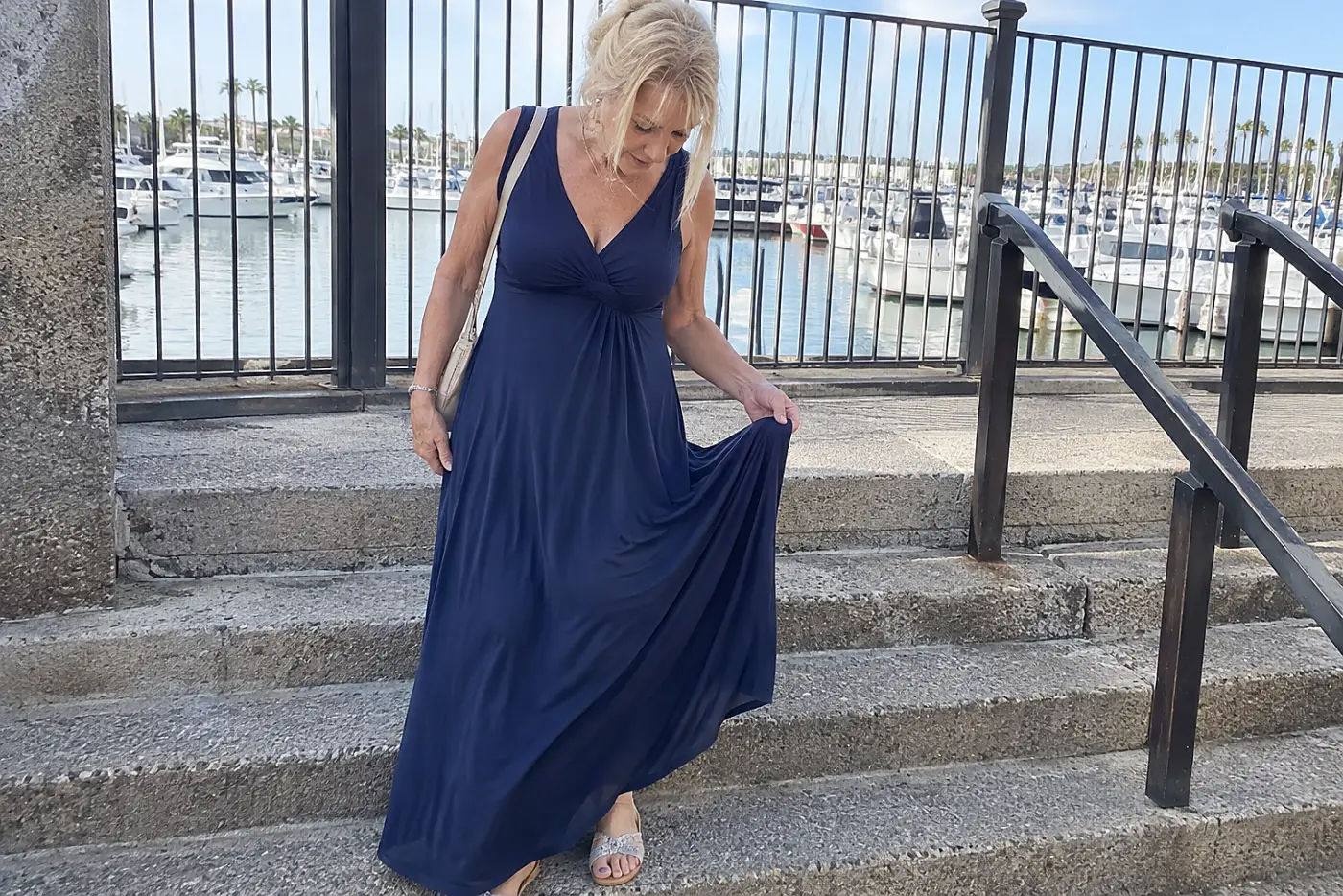 Older woman checking the hem of a maxi dress on outdoor steps near a harbor.
