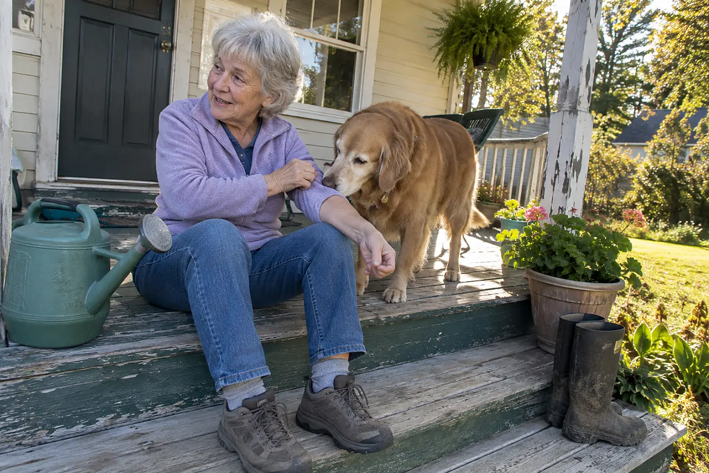 Older woman sitting on a porch step while her dog sniffs the same spot on her forearm.