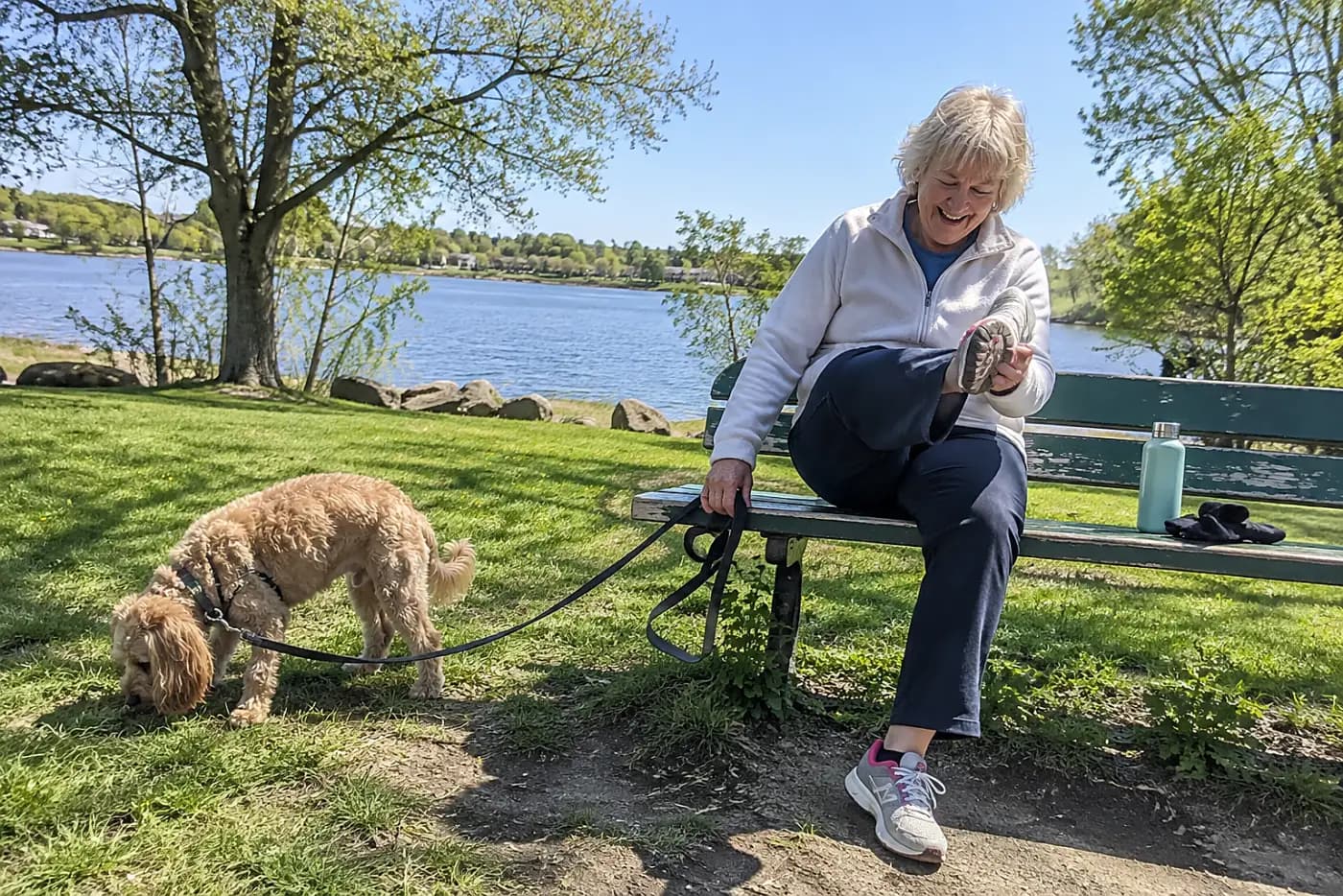 Older woman inspecting the sole of a shoe on a park bench while her dog sniffs nearby.