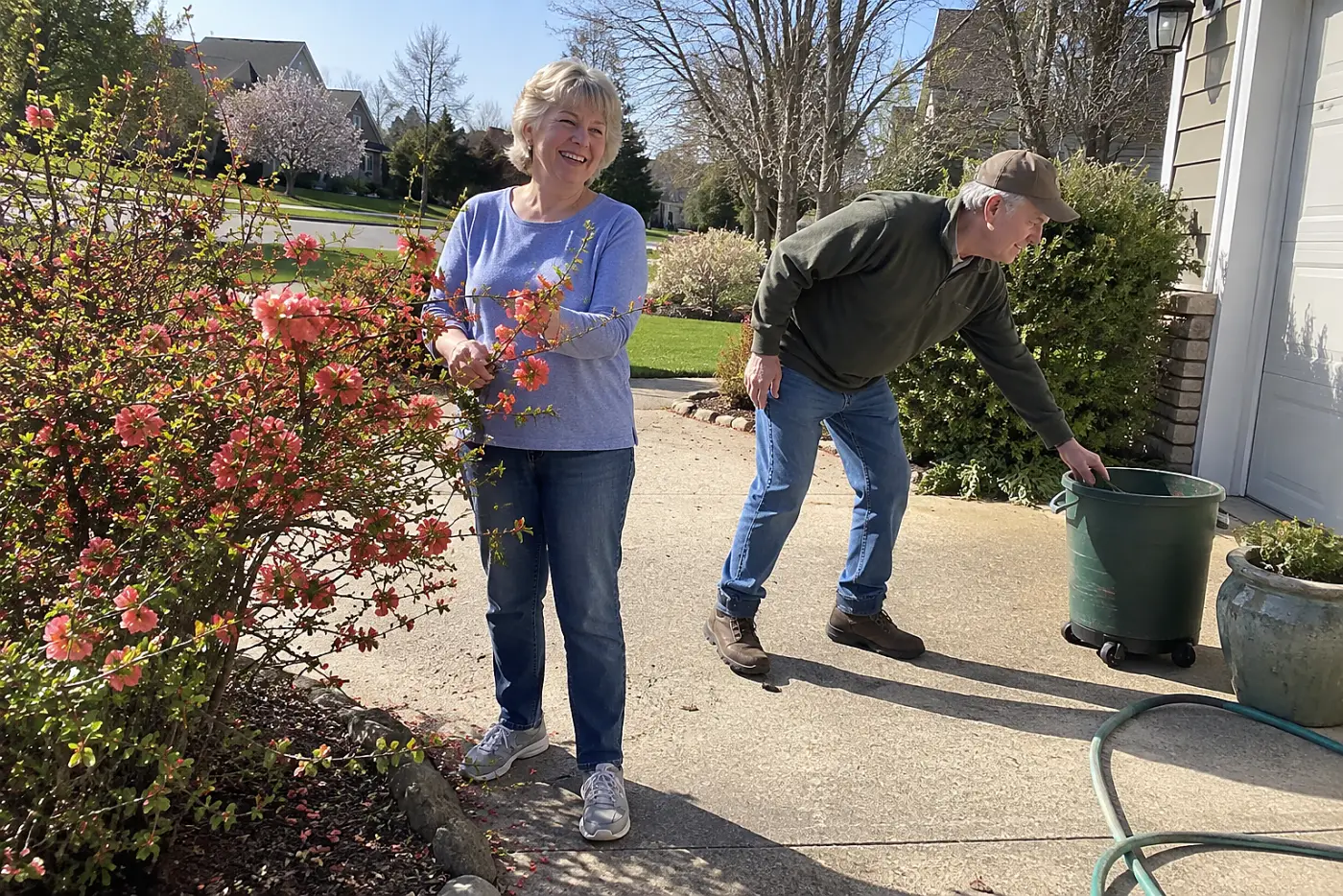A woman in her sixties smiles beside a coral flowering quince by the driveway.