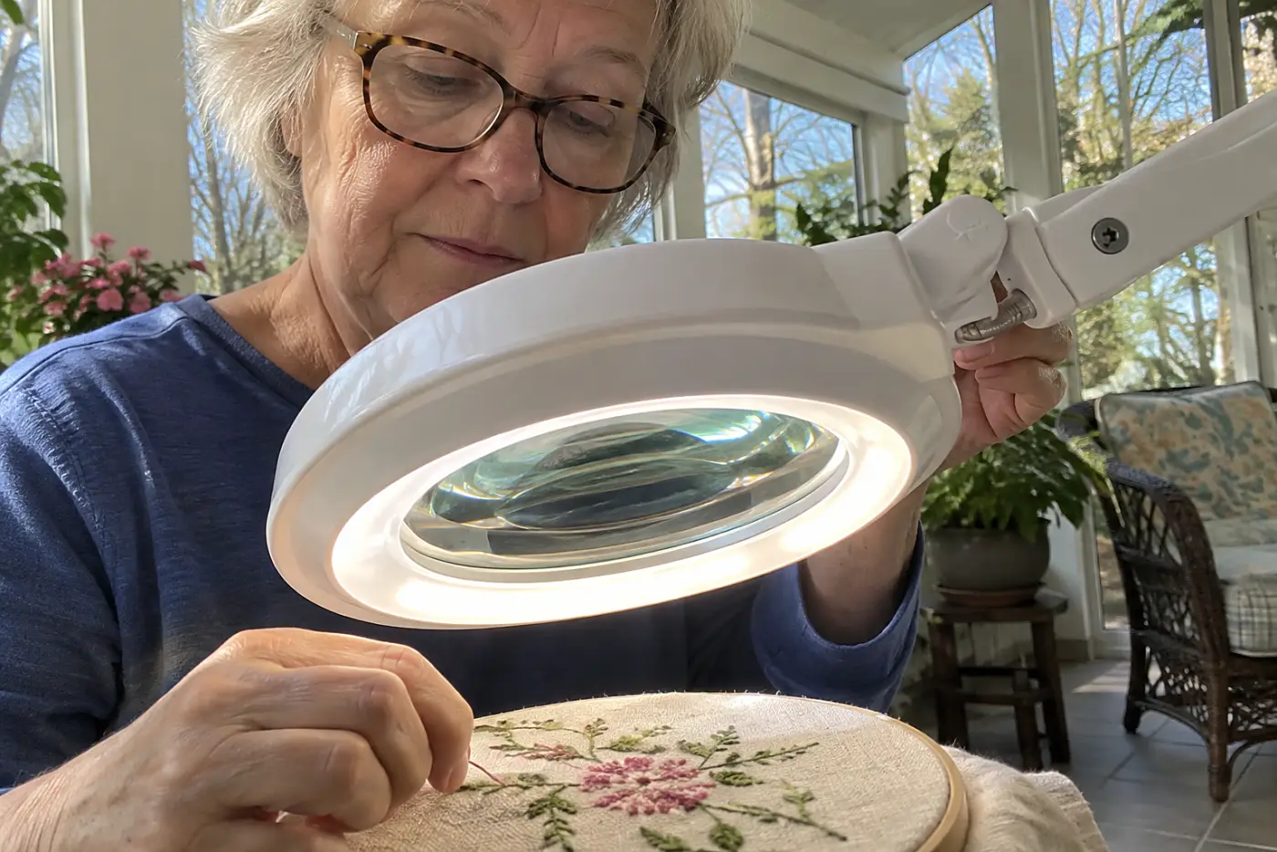 Woman in her sixties moving a magnifier light closer to her embroidery in a sunroom.