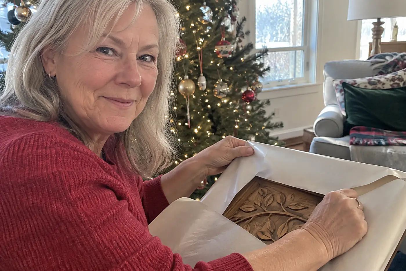 Woman in her sixties wrapping an engraved board beside a living room Christmas tree.