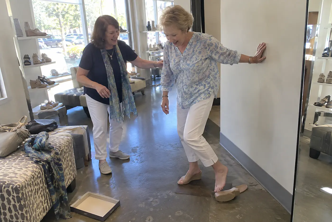 A low wedge mule fills the center of the frame on the foot of a woman in her sixties as she turns on a fitting-room floor, with no writing visible.