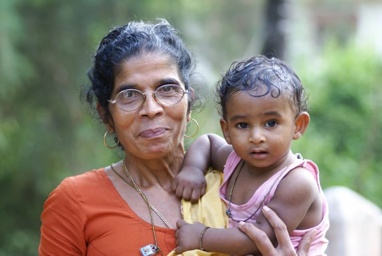 Smiling older Indian woman with glasses and hoop earrings holding a toddler outdoors against green foliage