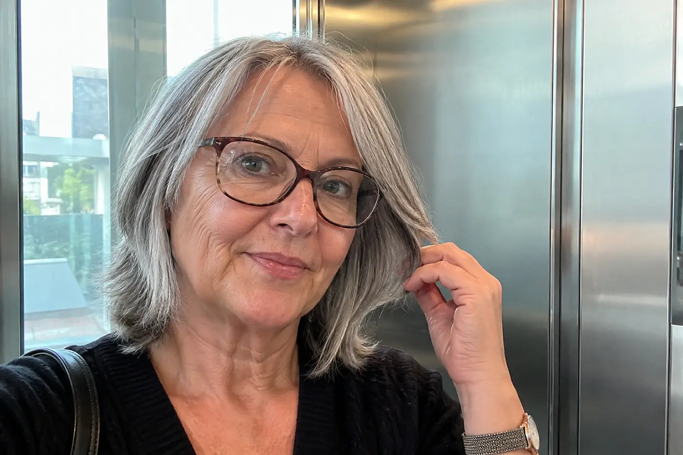 A woman with grey layered hair and glasses touches her hair inside an elevator.