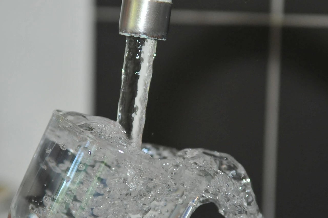 Water pours from a faucet into a clear glass, overflowing with splashing bubbles.