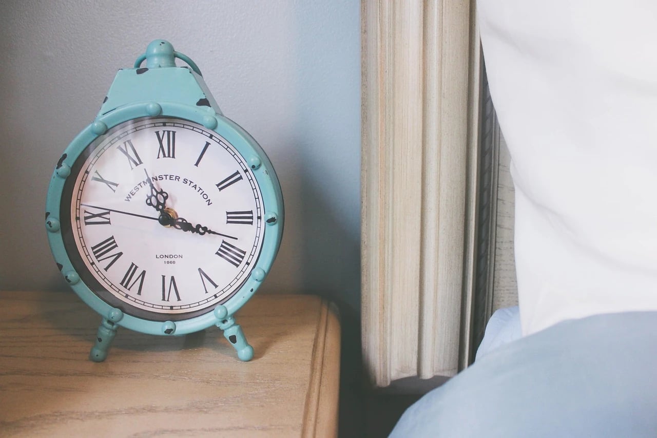 Teal vintage alarm clock with Roman numerals on a wooden nightstand next to a bed with white pillow