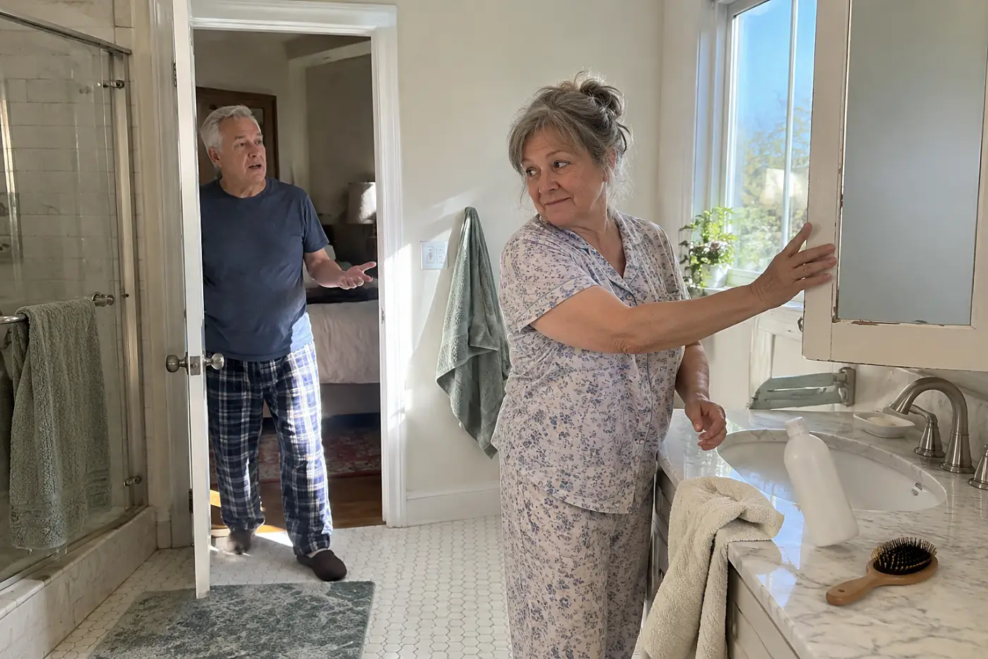 A woman in her sixties in floral pajamas closes a bathroom medicine cabinet mirror.