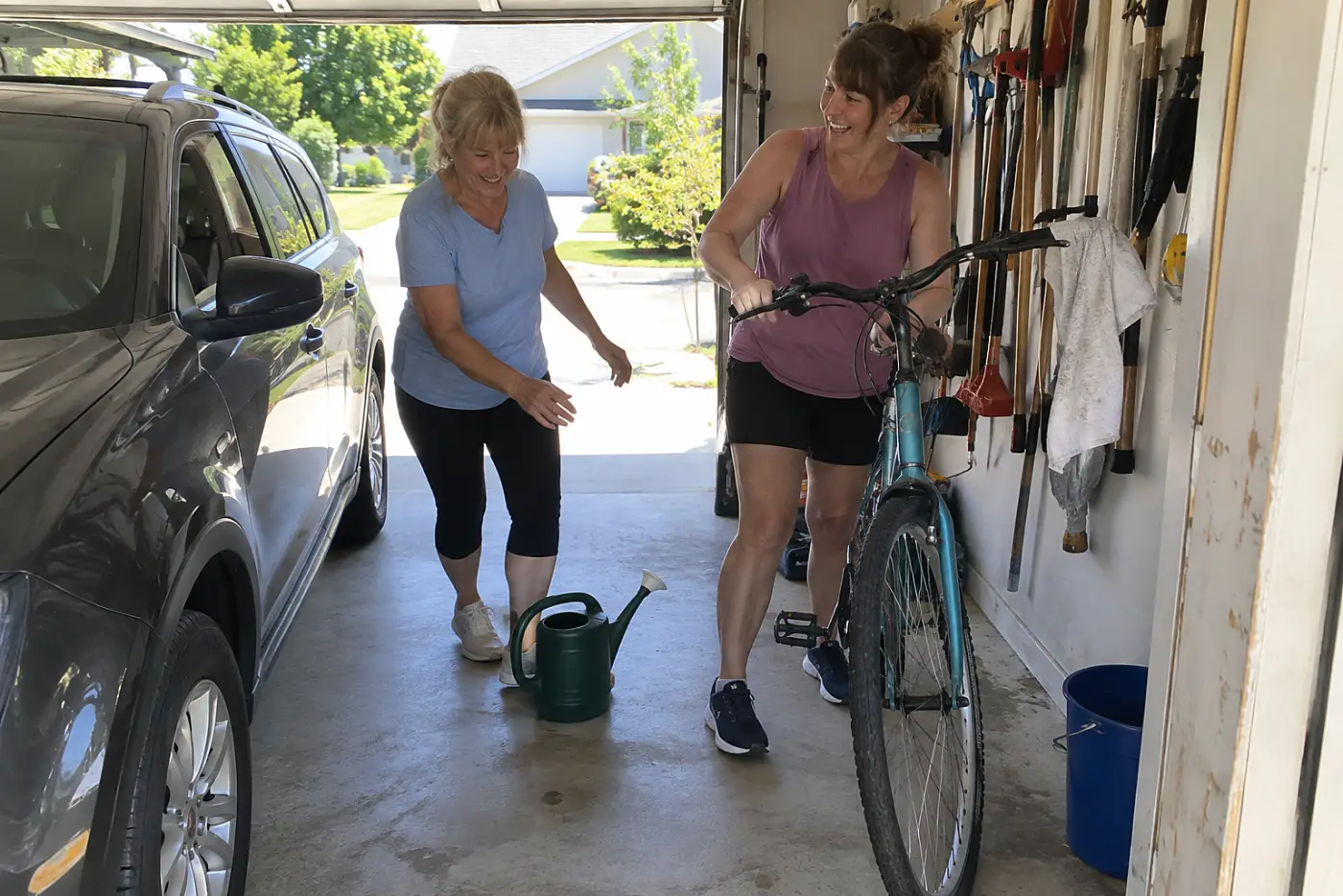 A woman in her fifties guides a bicycle past a parked car inside a garage.