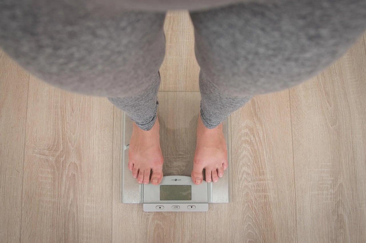 Overhead view of bare feet in gray leggings standing on a glass bathroom scale on wooden floor