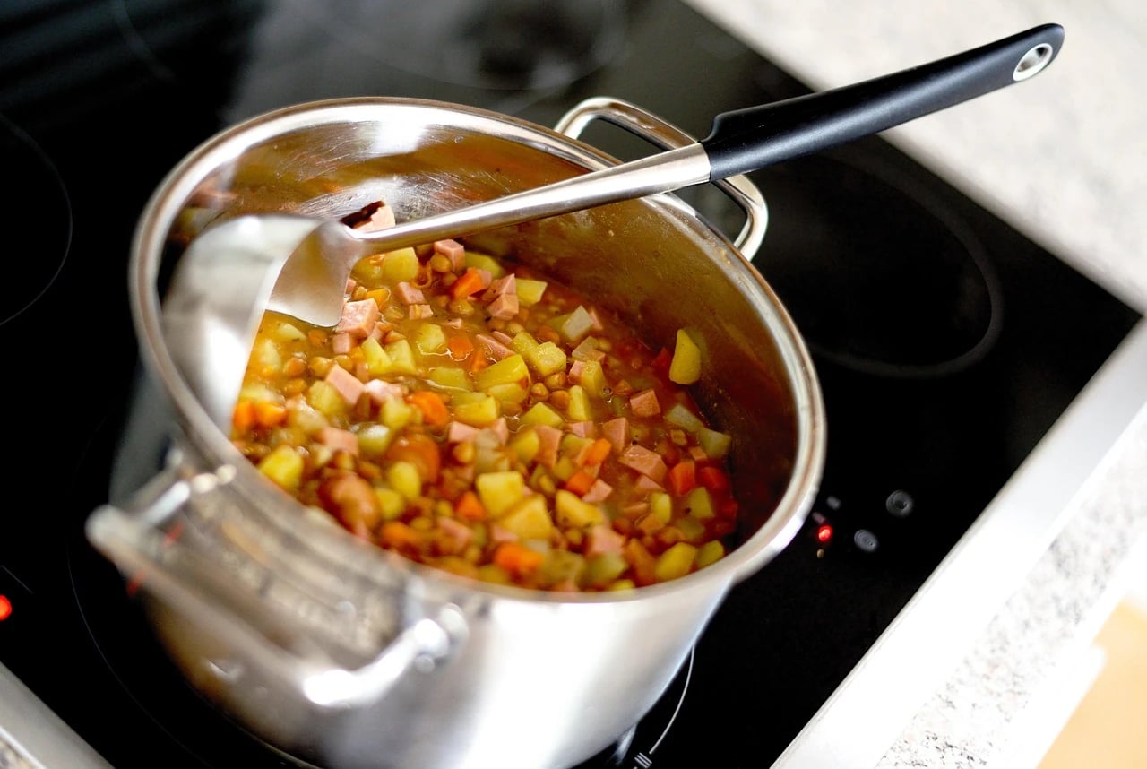 Stainless steel pot of lentil stew with diced vegetables and a ladle on a black cooktop