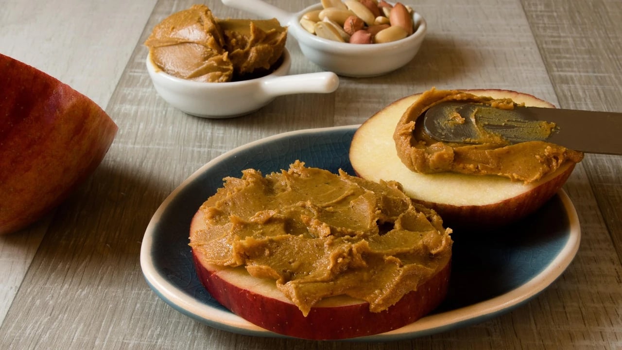 Two apple slices spread with peanut butter on a blue plate, with a knife and small bowls of peanuts