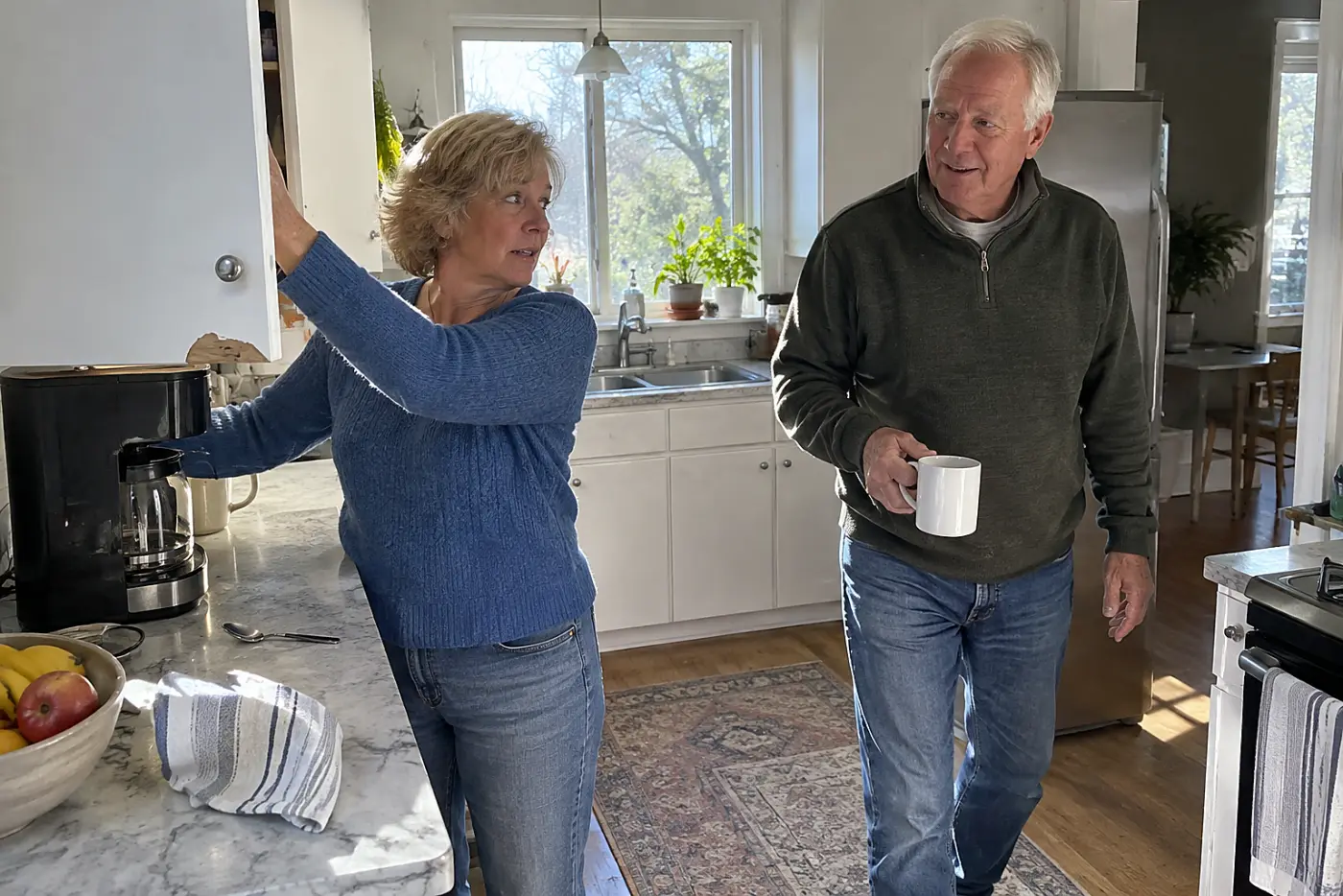 An older woman reaches toward a cupboard beside her coffee maker in the kitchen.
