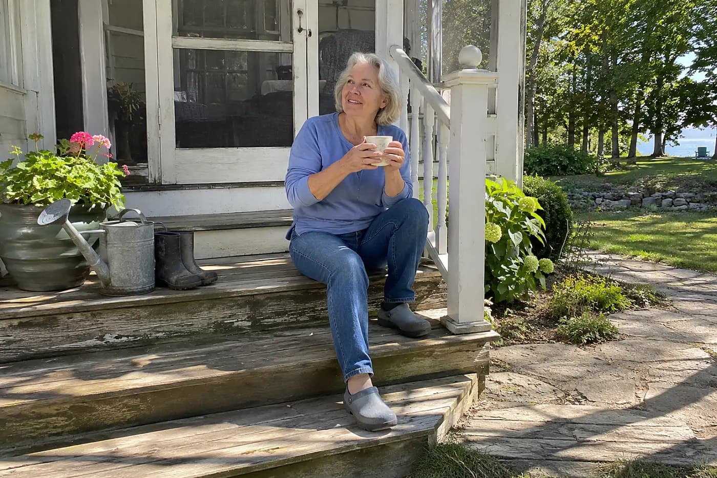An older woman sits alone on a porch step, holding a mug in both hands.