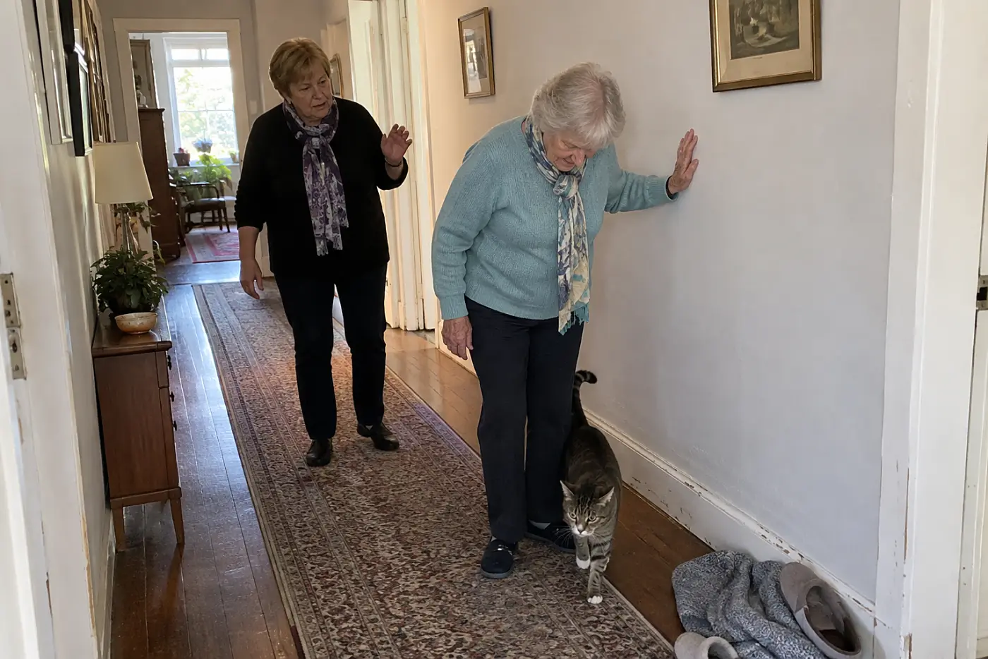 An older woman steadies herself against a hallway wall and looks down near her ankle.