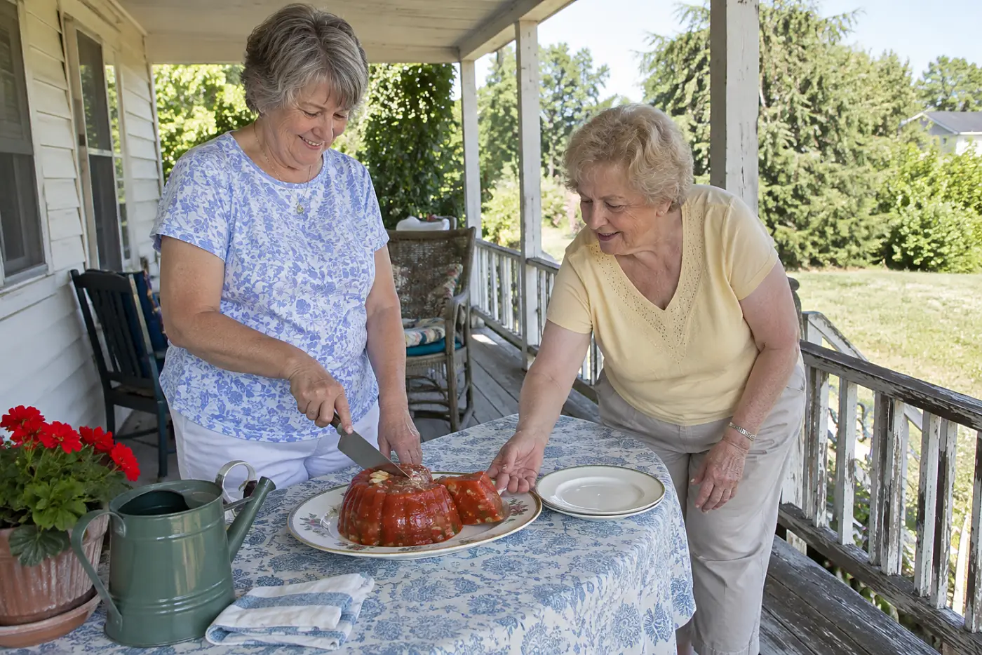 An older woman slices tomato aspic on a platter on a covered porch.