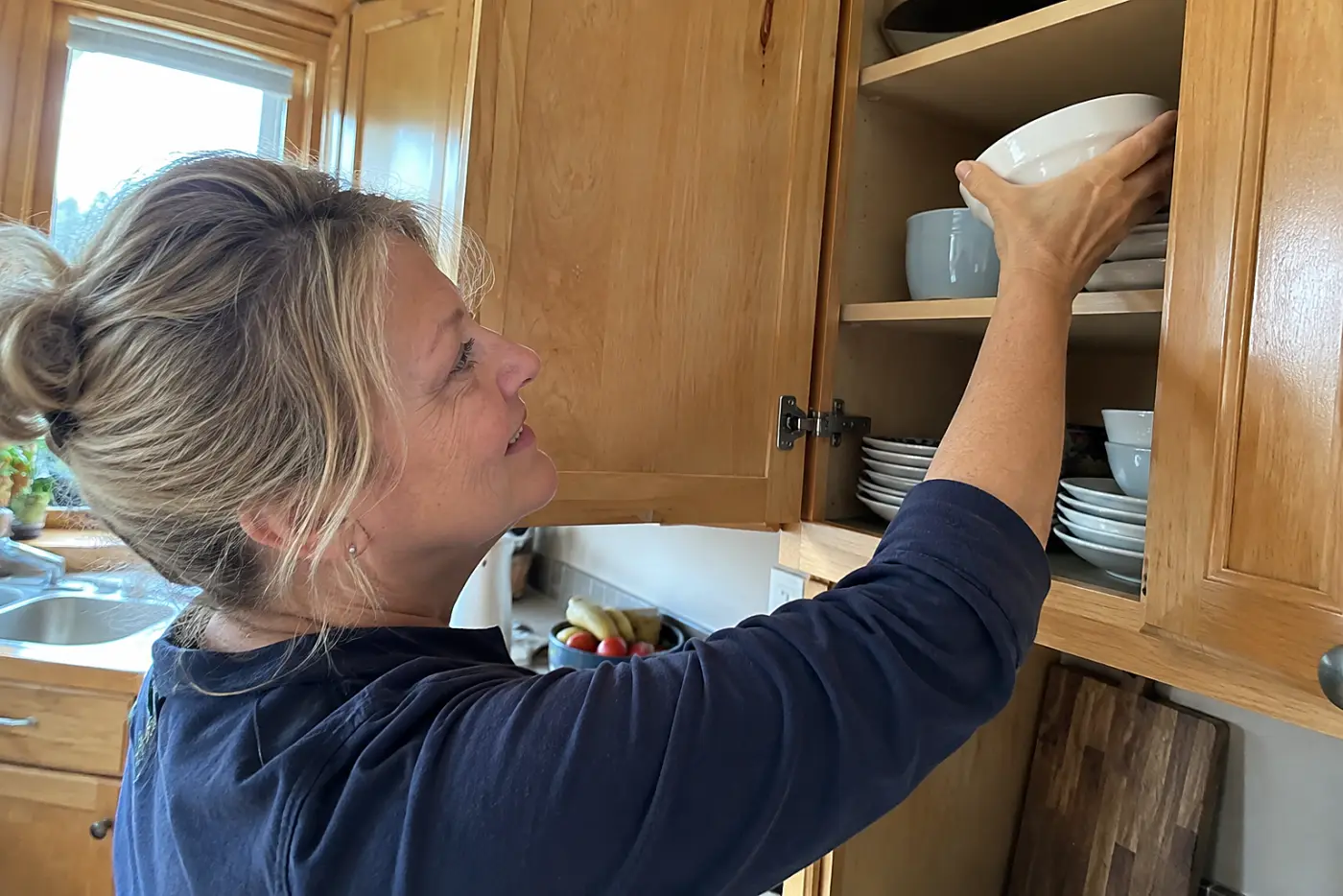 A woman over 50 puts a white bowl into a honey-oak kitchen cabinet.