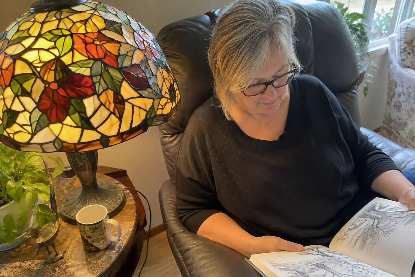 A woman in glasses reads a book in a recliner beside a glowing faux-Tiffany lamp.