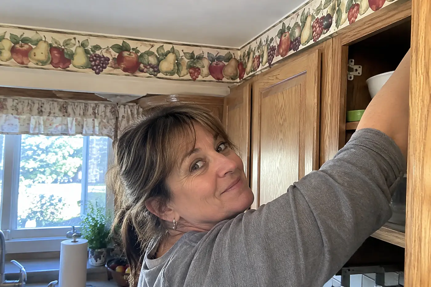 A woman in her fifties reaches into a kitchen cabinet beneath a fruit wallpaper border.
