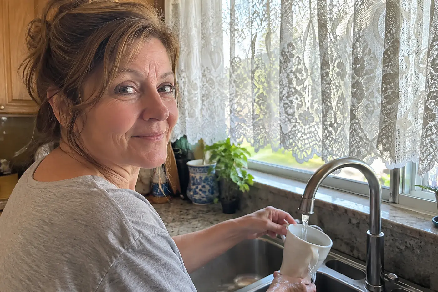 A smiling woman over 50 in a grey shirt fills a cup under lace curtains.
