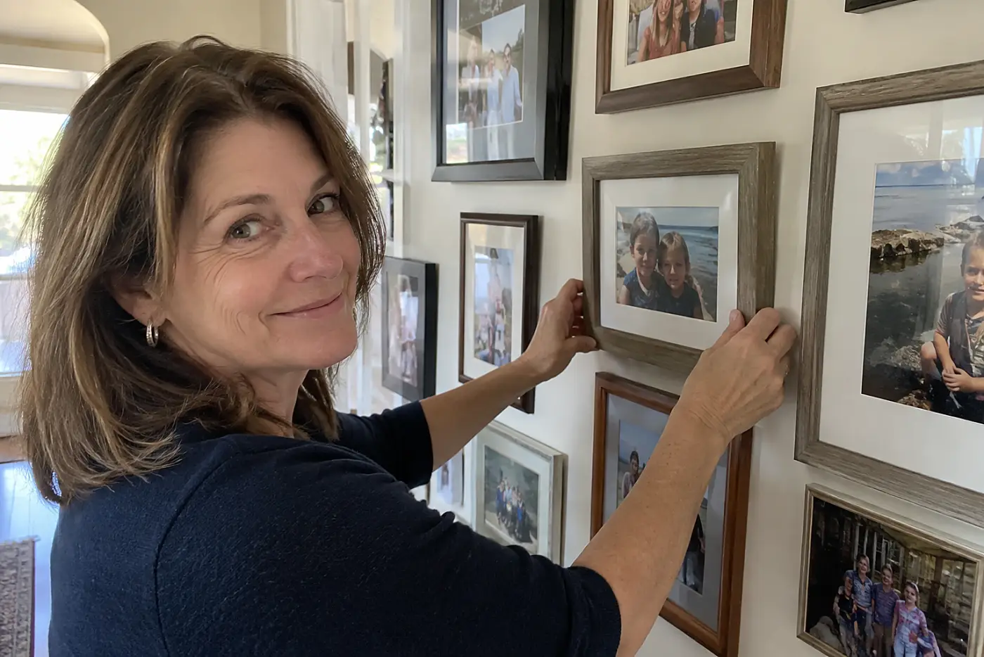 A woman over 50 straightens a picture frame on a wall filled with family portraits.