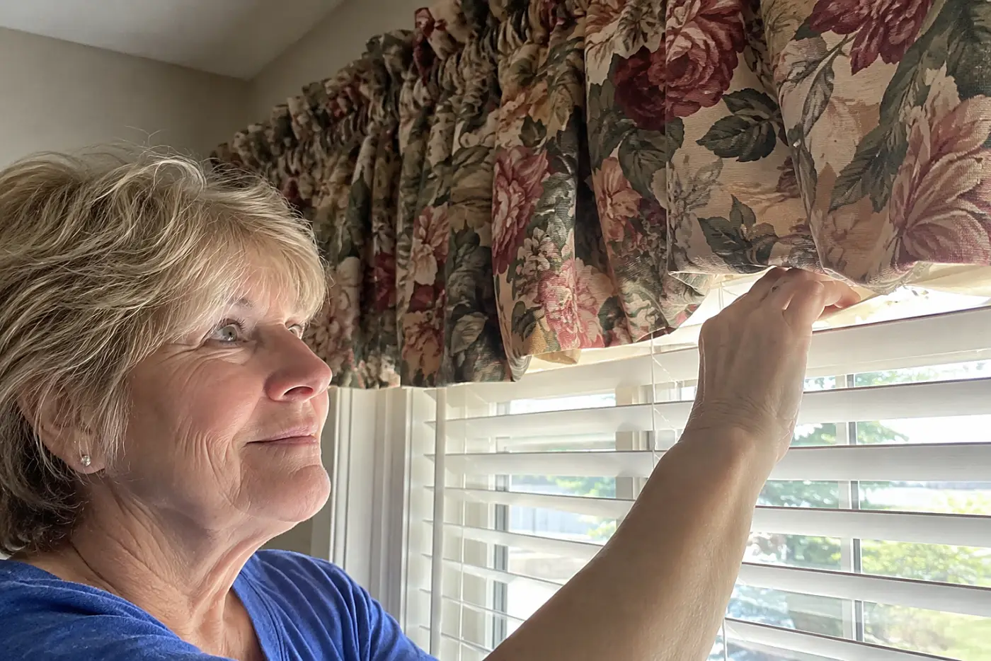 A woman in her sixties adjusts white window blinds beneath a heavy floral valance.