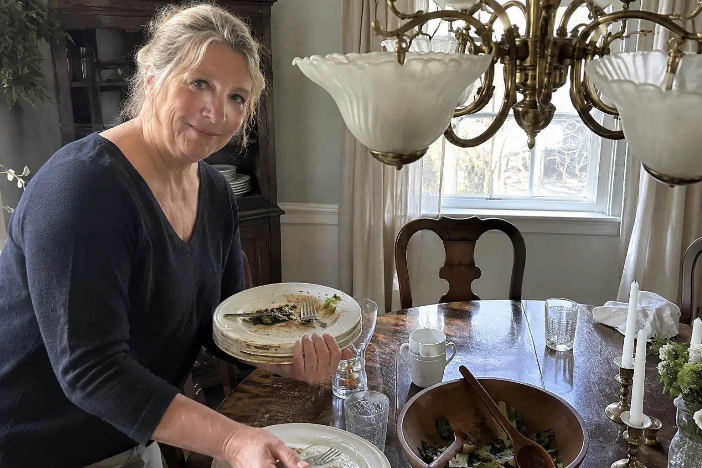 A woman in her sixties clears dirty plates from a table under a brass chandelier.