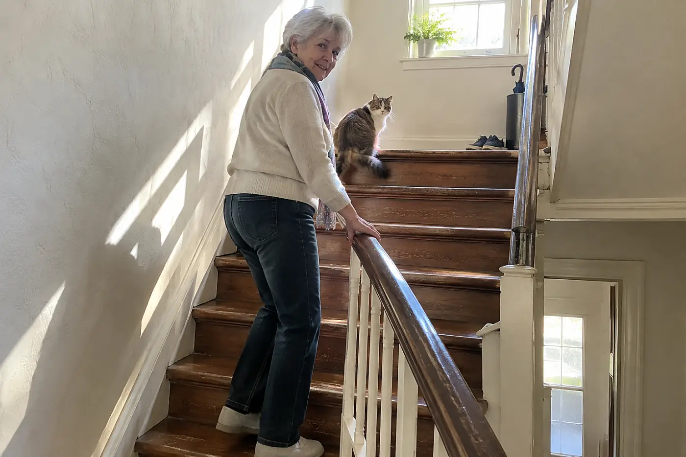 An older woman watching her cat pause on the stairs inside her home.