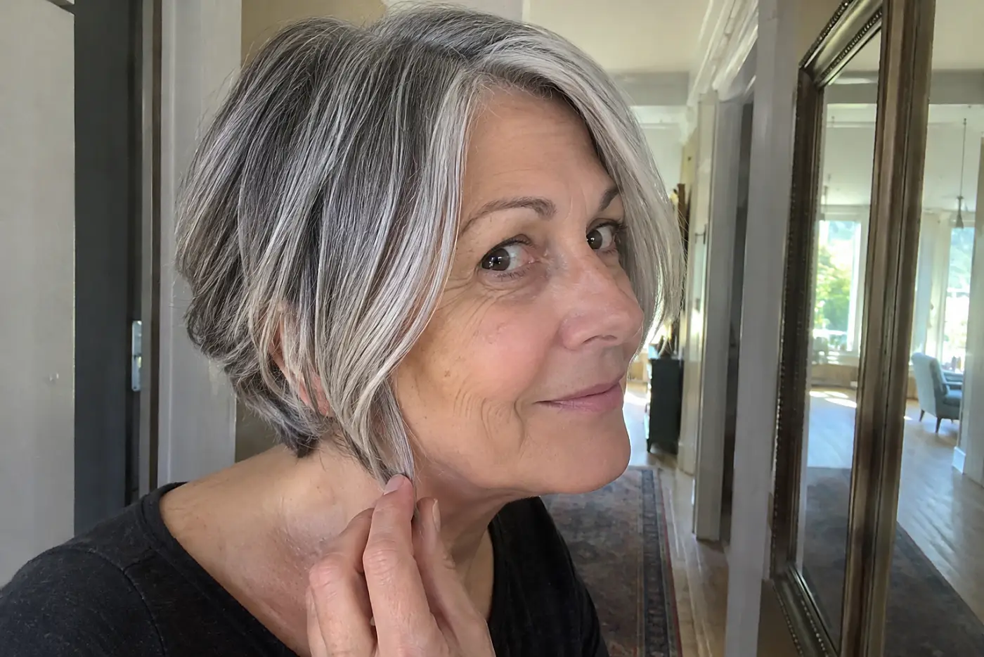An older woman looks at the short ends of her hair in a hallway mirror.