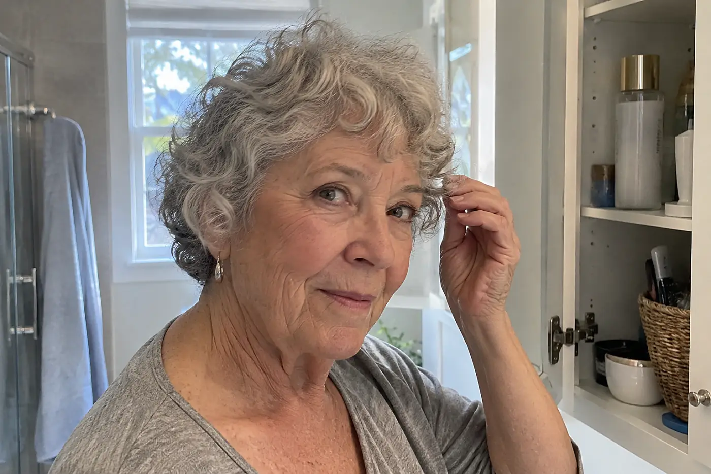 Woman arranging her cropped gray curls beside an open bathroom cabinet.