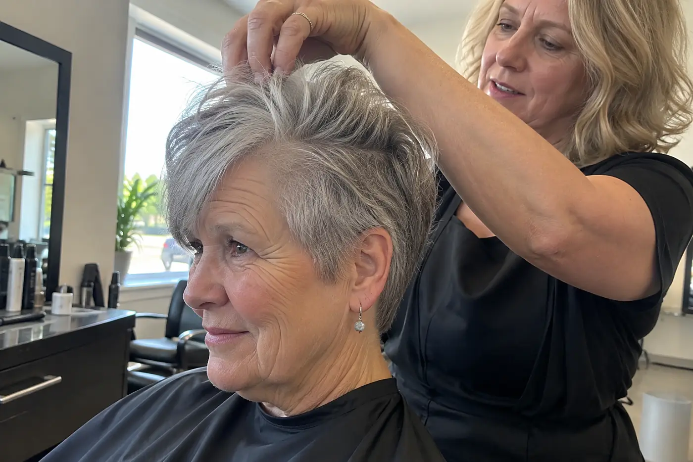Stylist lifting the top of a woman's short gray crop to show the hair beneath at a salon station.