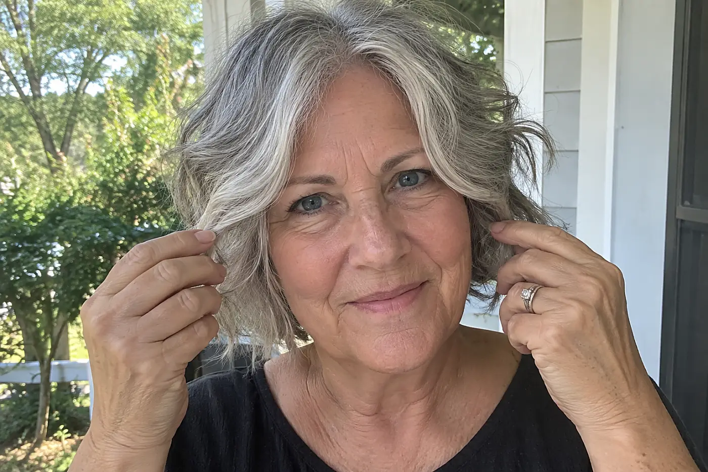 Woman separating the curtain bangs of her wavy gray bob on a front porch.