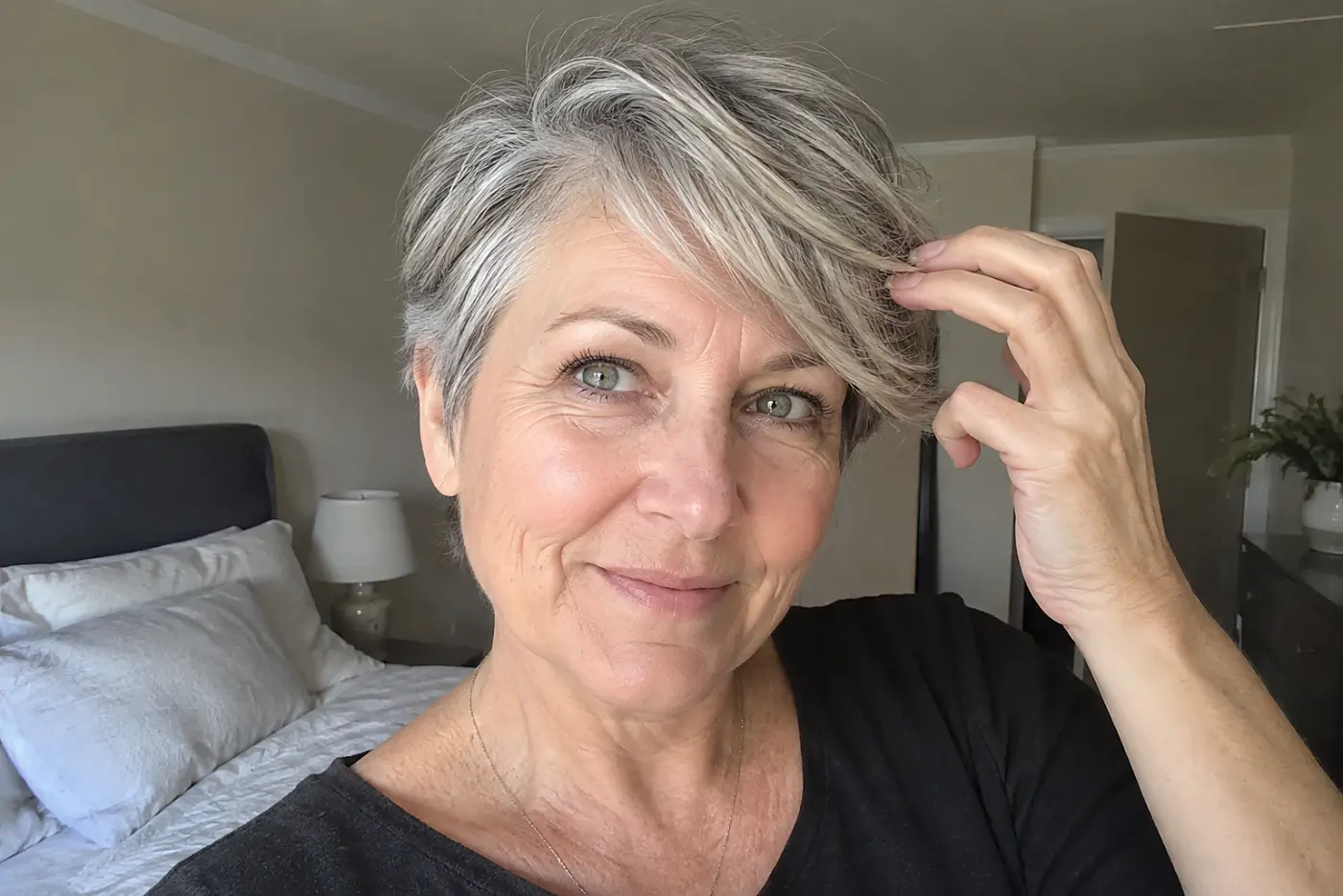Woman moving the side-swept bangs of her gray pixie with her fingers in a bedroom mirror.