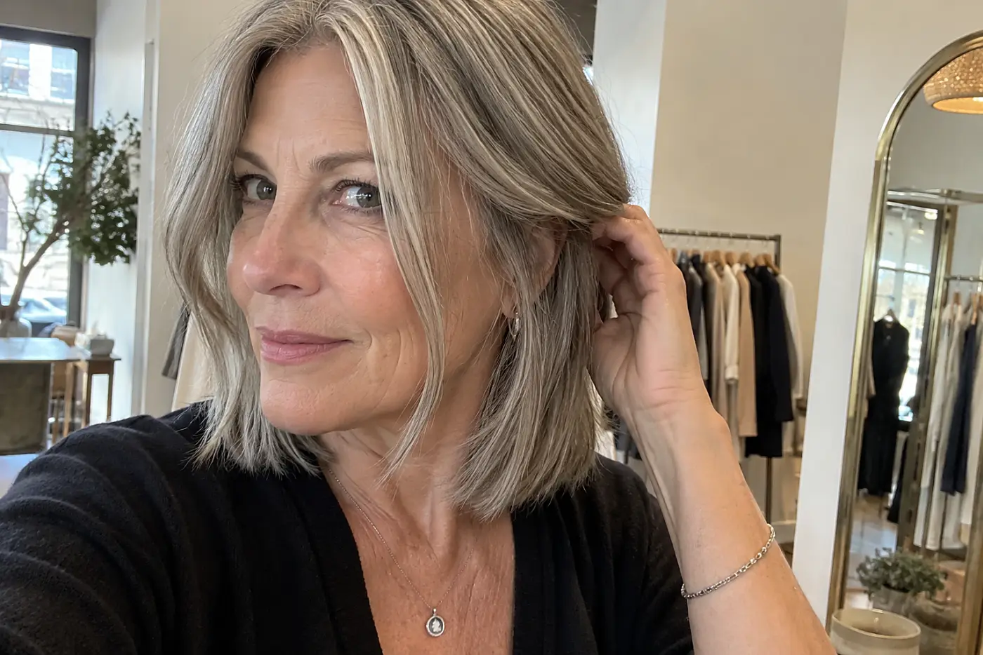Woman tucking her face-framing gray lob behind an ear in a boutique mirror.