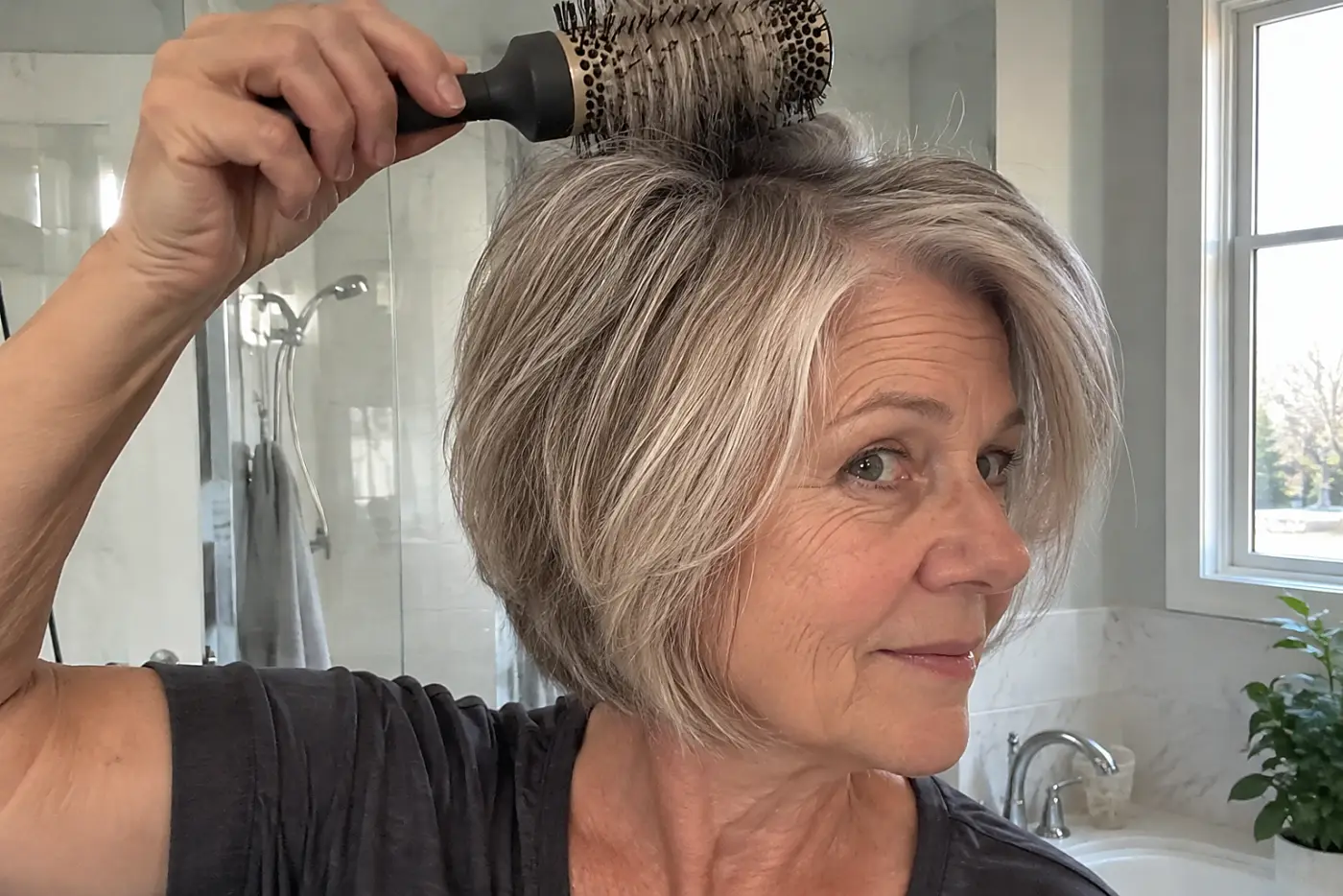 Woman lifting the roots of her gray bob with a round brush in a bathroom.
