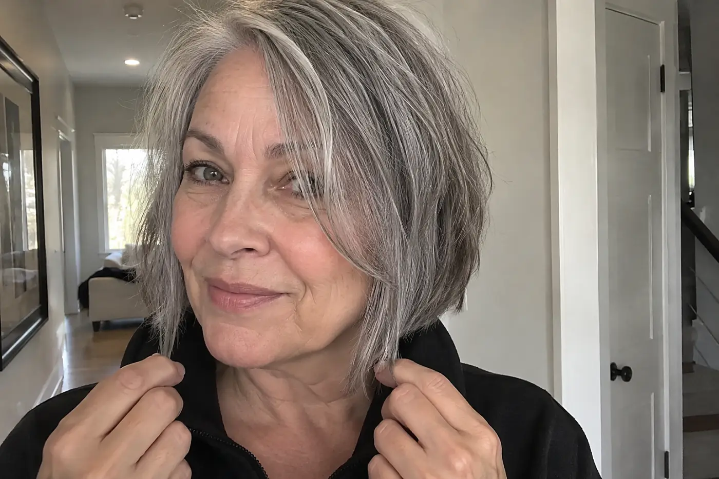 Woman holding the ends of her choppy gray bob against her dark jacket in a hallway.