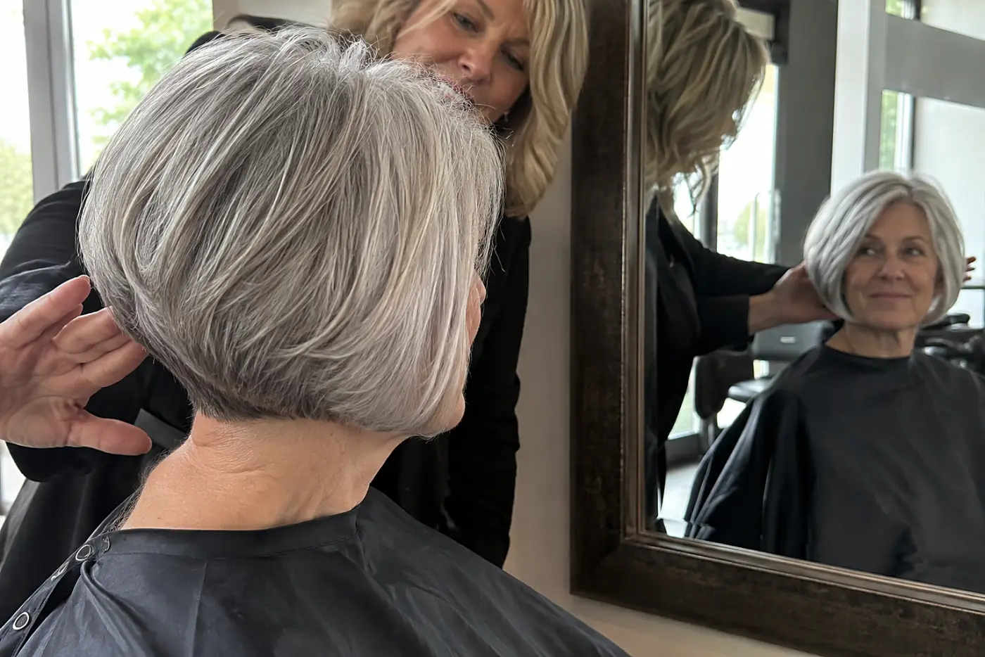 Stylist showing a woman the shorter back of her graduated gray bob in a salon mirror.