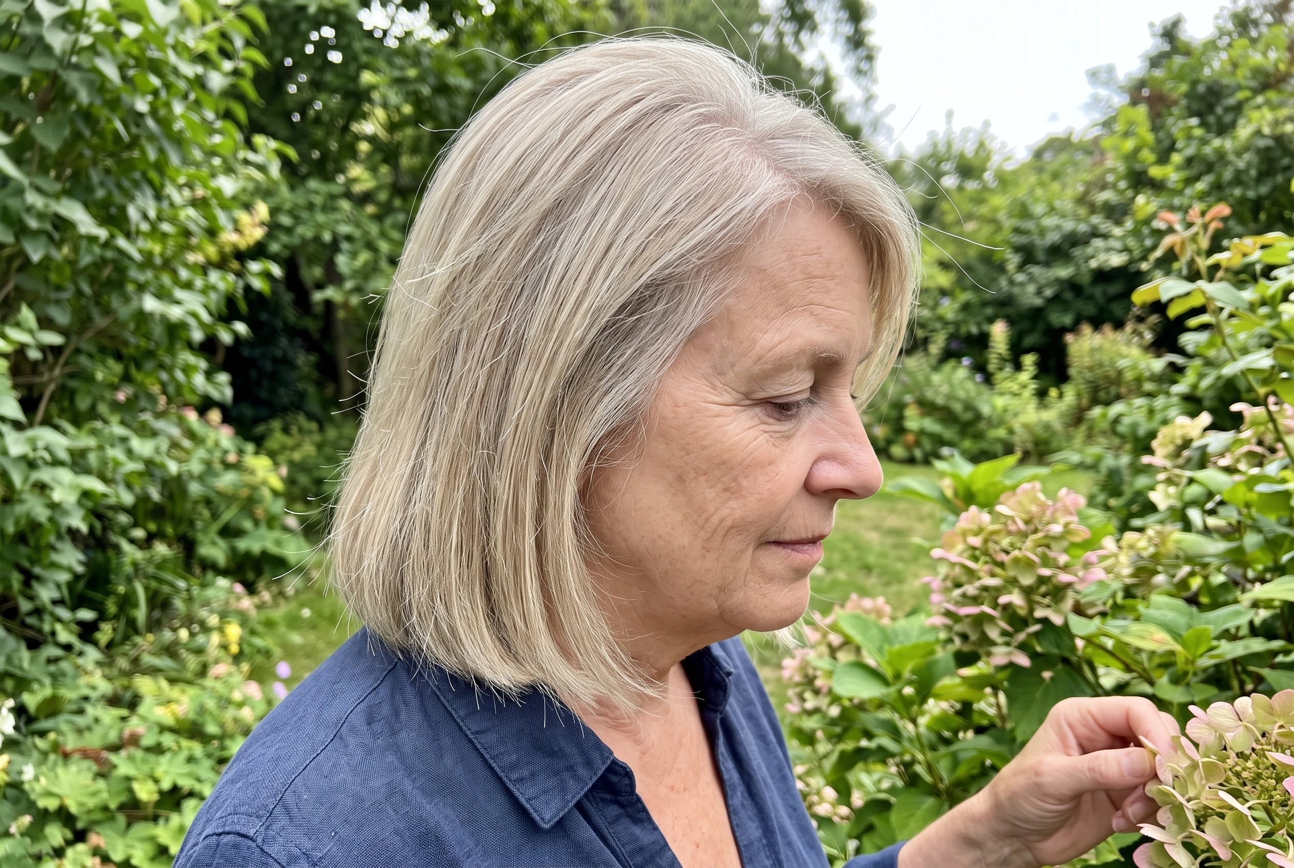 A woman in her sixties with shoulder-length ash blonde hair touches flowers in a garden.