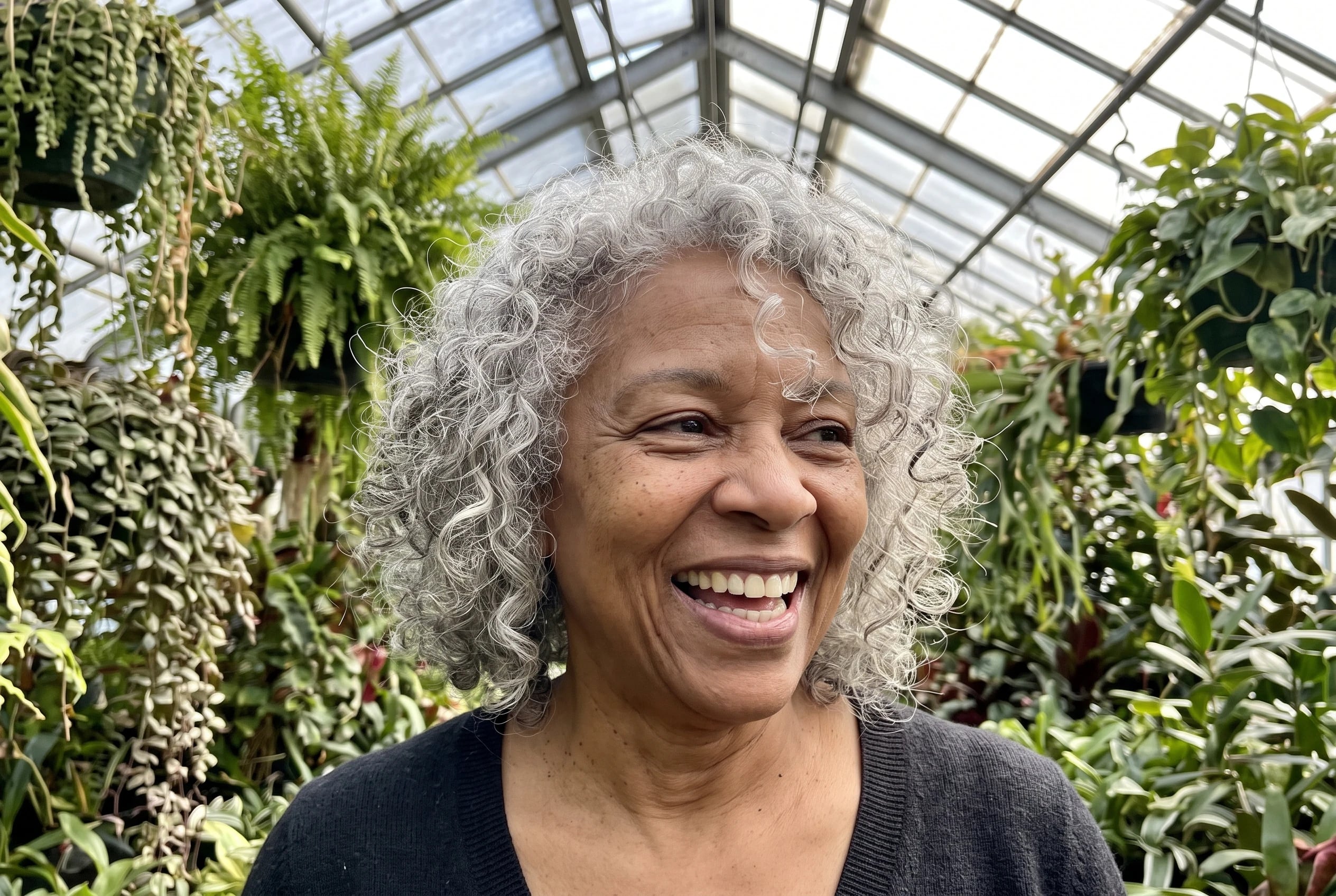 A smiling woman in her sixties with shoulder-length gray curls stands in a greenhouse.