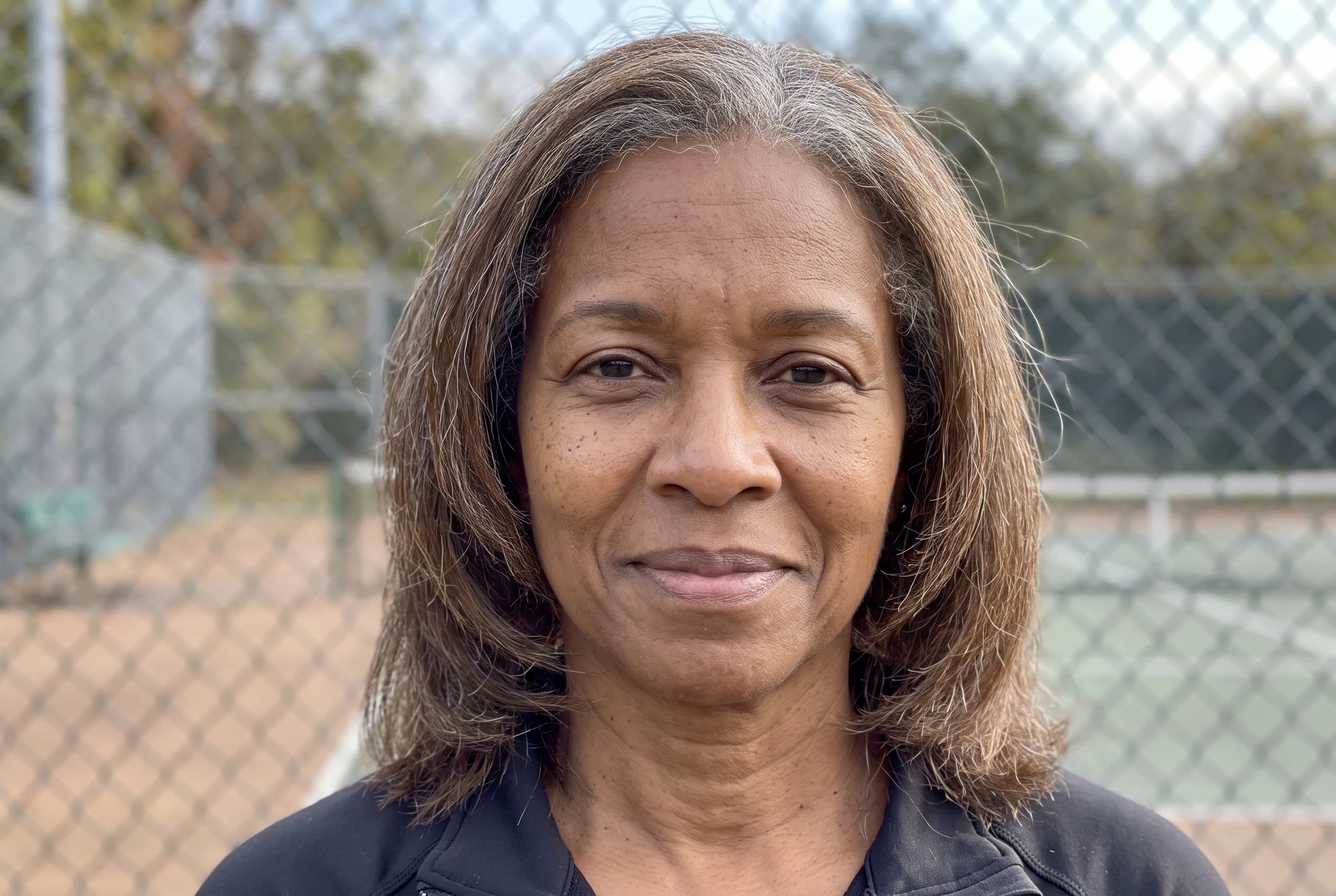 A woman in her sixties with layered, shoulder-length grey hair stands beside a tennis court.