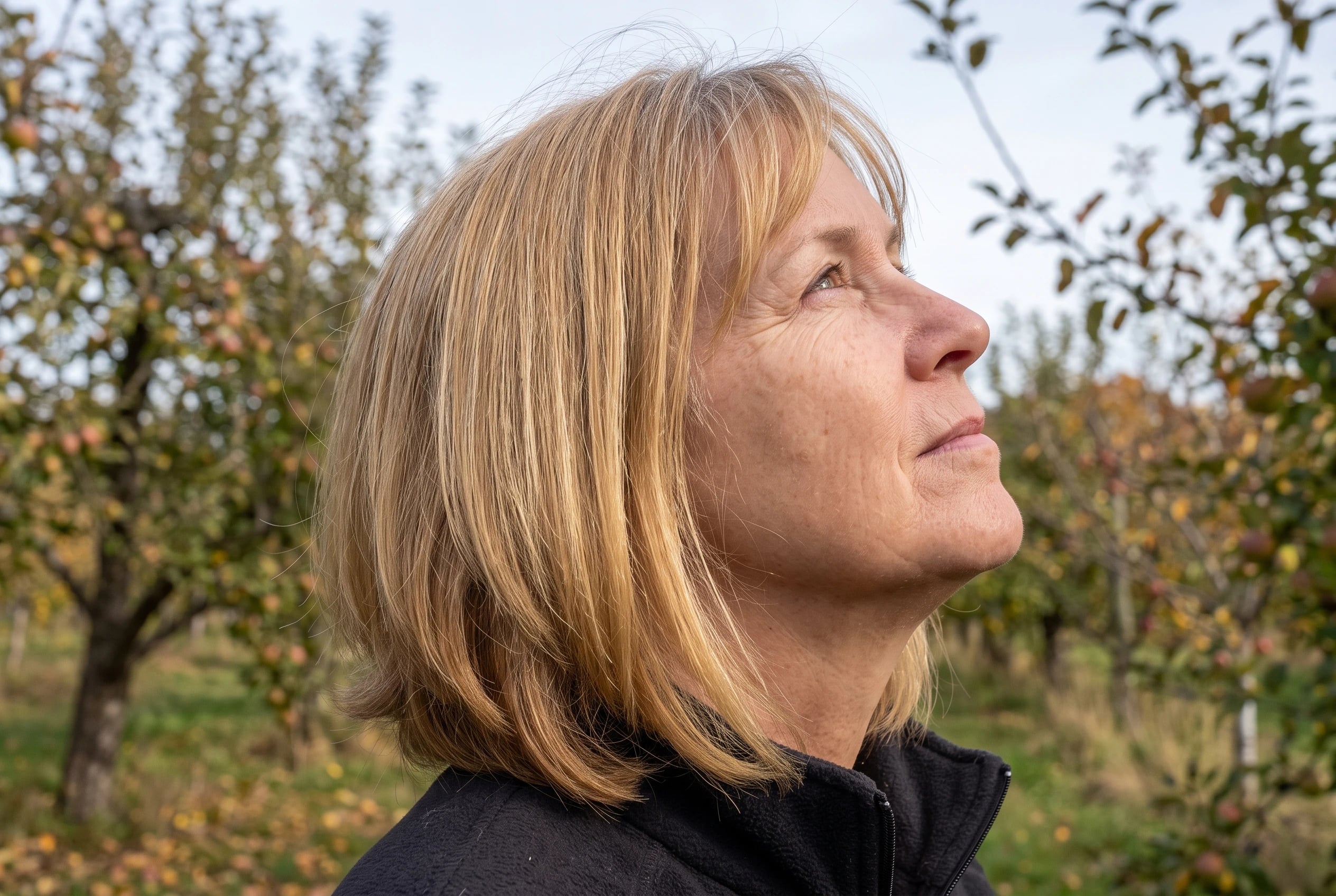 A woman in her sixties with shoulder-length golden blonde hair stands in an apple orchard.