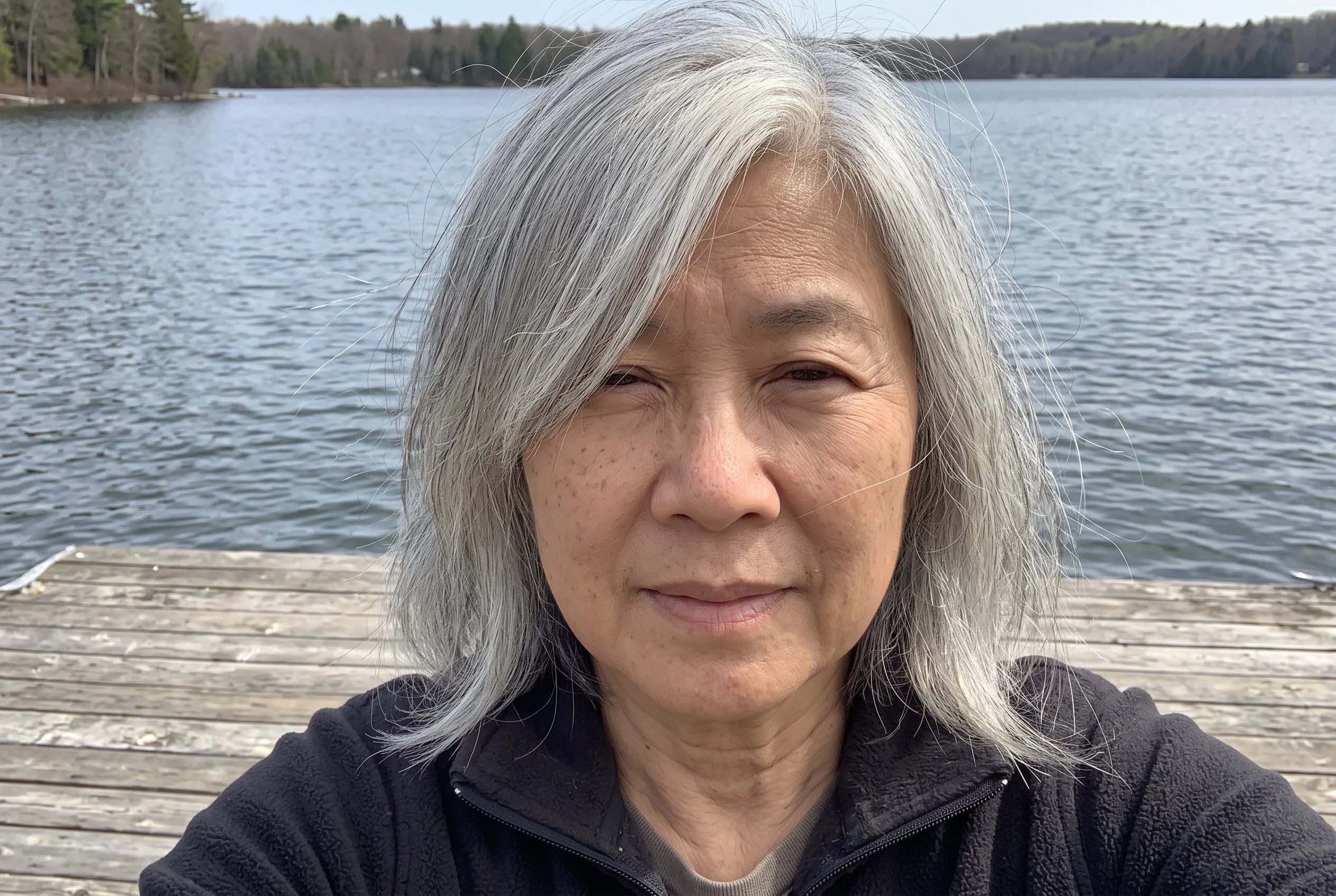 A woman in her sixties with deep parted silver hair stands on a wooden dock.