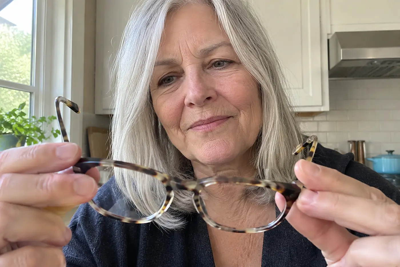 A woman in her sixties examines a pair of tortoiseshell glasses in her kitchen.