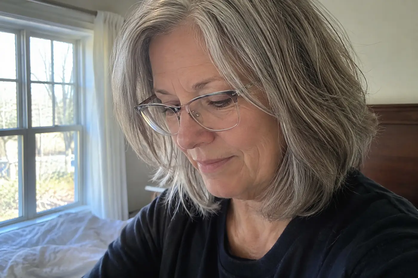 A woman in her sixties with shoulder-length silver hair wears silver rectangular glasses indoors.